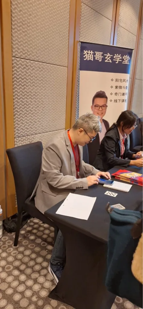 猫哥坐在“猫哥玄学堂”展位前，背景易拉宝显示提供阳宅风水，紫微斗数及奇门遁甲课程服务