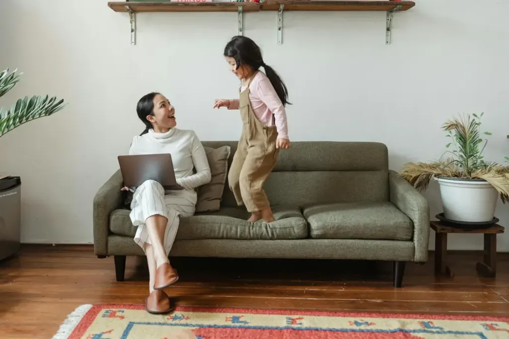 A happy Asian mother working on a laptop while her daughter plays on the sofa in a bright, clutter-free living room. This represents the ultimate goal of Feng Shui: creating a space with good Qi flow that supports both career success (Wealth) and family happiness (Harmony).