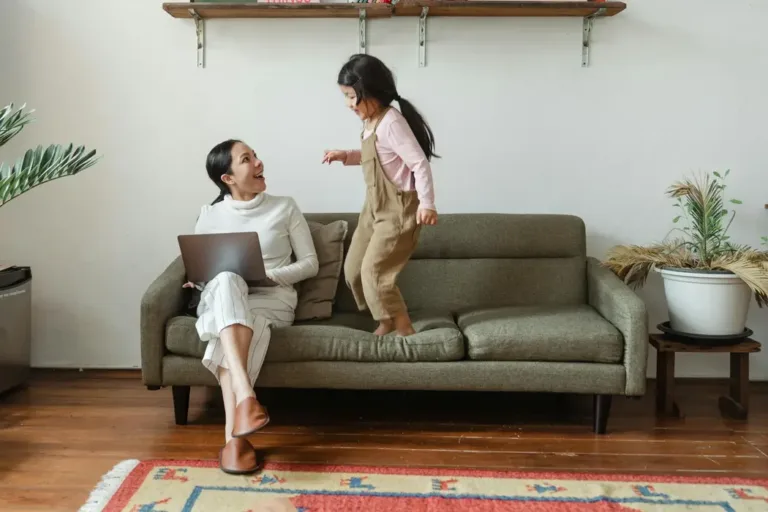 A happy Asian mother working on a laptop while her daughter plays on the sofa in a bright, clutter-free living room. This represents the ultimate goal of Feng Shui: creating a space with good Qi flow that supports both career success (Wealth) and family happiness (Harmony).