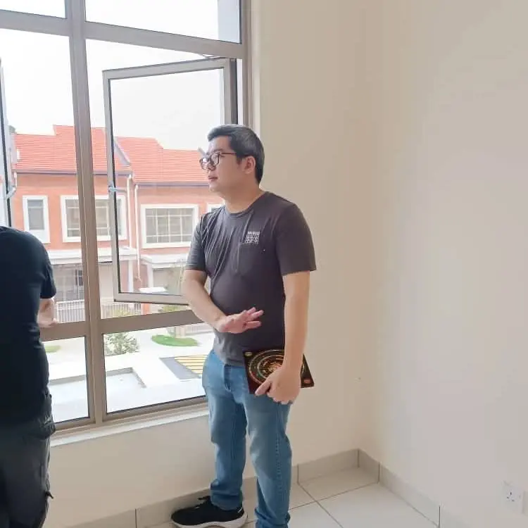 马来西亚风水师傅猫哥手持罗盘，站在新居窗前勘察阳宅风水的外围环境与气口。